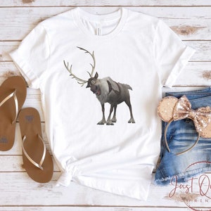 May include: White t-shirt with a cartoon image of a reindeer with antlers. The reindeer is brown and gray and has a surprised expression. The t-shirt is on a wooden surface with a pair of brown flip flops and a pair of denim shorts with a gold bow headband.