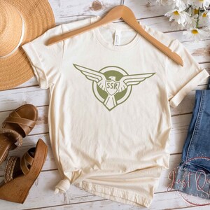 Op de afbeelding: Een beige T-shirt met een groene adelaar afbeelding en de tekst "SSR" in een cirkel. Het shirt ligt op een houten oppervlak met een strohoed, sandalen en een blauwe jeans.