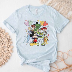 Puede incluir: Camiseta azul claro con un estampado gr&aacute;fico de Mickey Mouse, Woody, Buzz Lightyear, Jessie, Rex y Slinky Dog. Los personajes est&aacute;n rodeados de globos y estrellas de colores.