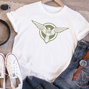 Op de afbeelding: Wit T-shirt met een groene adelaar afbeelding en de tekst "SSR" in een cirkel.