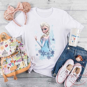 May include: A white t-shirt featuring a graphic of Elsa from Frozen, paired with blue jeans, white sneakers, and a Disney-themed backpack. A rose gold sequined Minnie Mouse ear headband and a Disney-themed tumbler are also present.