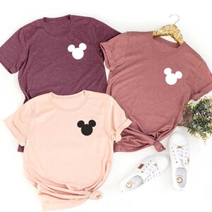 K&ouml;nnte beinhalten: Drei T-Shirts mit einer Mickey-Maus-Silhouette auf der Brusttasche. Die Shirts sind in den Farben Rosa und Rotbraun.