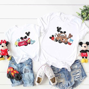 May include: Two white t-shirts with a cartoon image of a red car and a brown truck with Mickey Mouse ears and balloons. The shirts are paired with denim shorts and sneakers.
