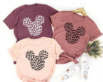 May include: Three short-sleeved t-shirts in burgundy, mauve, and blush pink, each featuring a leopard print Mickey Mouse head design. The shirts are displayed with white sneakers and a gold hanger.