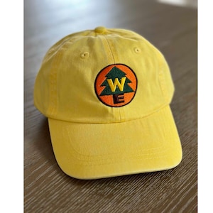 May include: A yellow baseball cap with a green pine tree and the letters "W" and "E" embroidered in orange and brown on a circular orange patch.