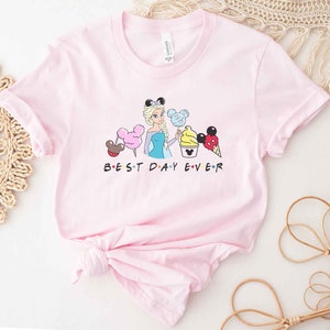 May include: Pink t-shirt with a graphic of a Disney princess character holding a Mickey Mouse ice cream cone and other Disney-themed treats. The text "BEST DAY EVER" is printed below the graphic.