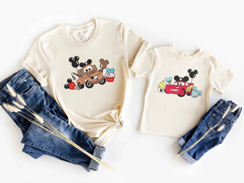 May include: Two beige t-shirts with a Disney Cars theme. The larger shirt features Mater with Mickey Mouse ears and balloons. The smaller shirt features Lightning McQueen with Mickey Mouse ears and balloons. Both shirts have a Mickey Mouse ice cream cone and cupcake.