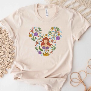 Puede incluir: Una camiseta beige con una silueta de Mickey Mouse compuesta por varias ilustraciones con tem&aacute;tica marina, incluyendo a Ariel de La Sirenita, conchas marinas, estrellas de mar y algas.