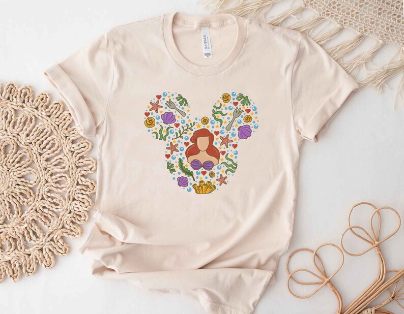 Puede incluir: Una camiseta beige con una silueta de Mickey Mouse compuesta por varias ilustraciones con tem&aacute;tica marina, incluyendo a Ariel de La Sirenita, conchas marinas, estrellas de mar y algas.