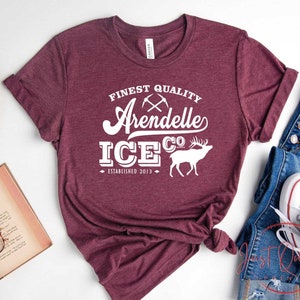 Puede incluir: Camiseta color burdeos con texto blanco que dice "Finest Quality Arendelle Ice Co. Established 2013". El texto est&aacute; en una fuente de estilo vintage e incluye un gr&aacute;fico de picos cruzados y un alce estilizado.