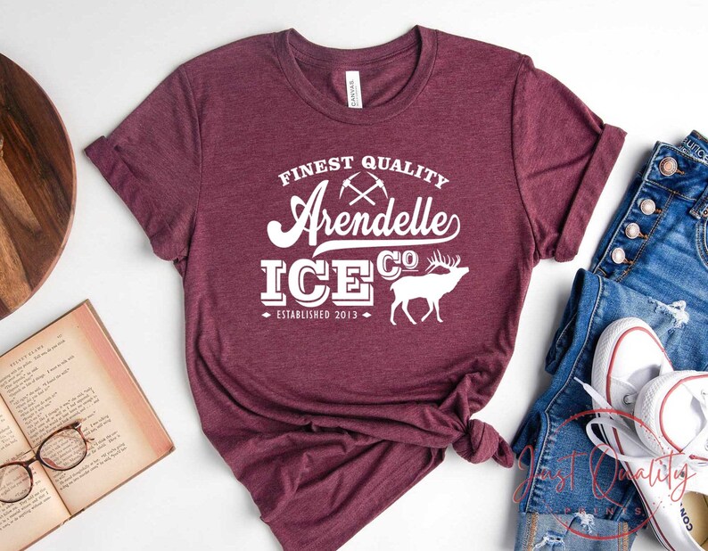 Puede incluir: Camiseta color burdeos con texto blanco que dice "Finest Quality Arendelle Ice Co. Established 2013". El texto est&aacute; en una fuente de estilo vintage e incluye un gr&aacute;fico de picos cruzados y un alce estilizado.