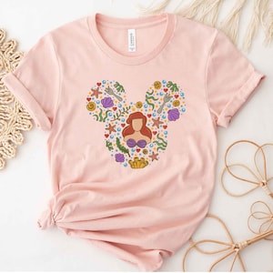 Puede incluir: Camiseta rosa con una silueta de Mickey Mouse compuesta por varias im&aacute;genes de La Sirenita, incluyendo a Ariel, conchas marinas y algas.