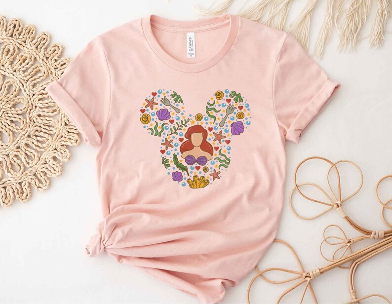 Puede incluir: Camiseta rosa con una silueta de Mickey Mouse compuesta por varias im&aacute;genes de La Sirenita, incluyendo a Ariel, conchas marinas y algas.