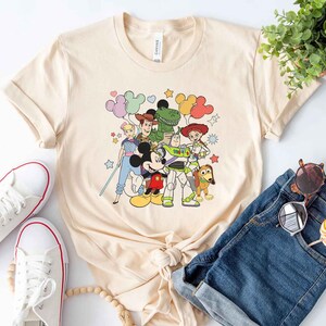 Puede incluir: Una camiseta de color beige claro con un estampado gr&aacute;fico de Mickey Mouse, Woody, Buzz Lightyear, Jessie, Rex y un extraterrestre verde de Toy Story. Los personajes est&aacute;n rodeados de globos de colores y estrellas.
