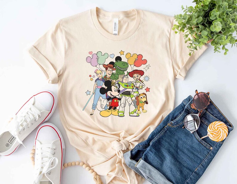 Puede incluir: Una camiseta de color beige claro con un estampado gr&aacute;fico de Mickey Mouse, Woody, Buzz Lightyear, Jessie, Rex y un extraterrestre verde de Toy Story. Los personajes est&aacute;n rodeados de globos de colores y estrellas.