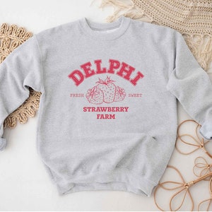 Op de afbeelding: Grijs sweatshirt met een rood grafisch ontwerp met de tekst "Delphi Strawberry Farm" met een tekening van aardbeien. De tekst zegt "Fresh" en "Sweet" aan weerszijden van de aardbeien.