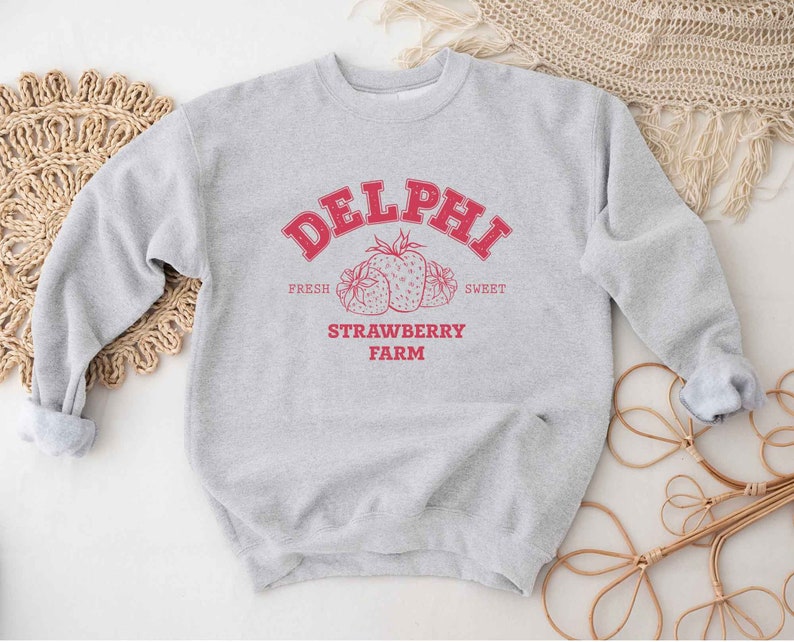 Op de afbeelding: Grijs sweatshirt met een rood grafisch ontwerp met de tekst "Delphi Strawberry Farm" met een tekening van aardbeien. De tekst zegt "Fresh" en "Sweet" aan weerszijden van de aardbeien.
