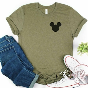 K&ouml;nnte beinhalten: Ein gr&uuml;nes T-Shirt mit einer schwarzen Mickey-Maus-Silhouette auf der Vorderseite. Das Shirt ist gefaltet und liegt auf einer wei&szlig;en Oberfl&auml;che mit einem Paar wei&szlig;en Sneakers und einer blauen Jeans.