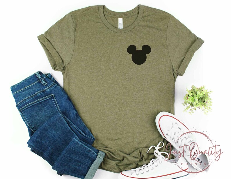 K&ouml;nnte beinhalten: Ein gr&uuml;nes T-Shirt mit einer schwarzen Mickey-Maus-Silhouette auf der Vorderseite. Das Shirt ist gefaltet und liegt auf einer wei&szlig;en Oberfl&auml;che mit einem Paar wei&szlig;en Sneakers und einer blauen Jeans.