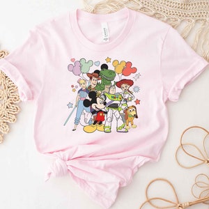 Puede incluir: Camiseta rosa con un estampado gr&aacute;fico de Mickey Mouse, Woody, Buzz Lightyear, Jessie, Rex y un extraterrestre verde de Toy Story. Los personajes est&aacute;n rodeados de estrellas y globos.