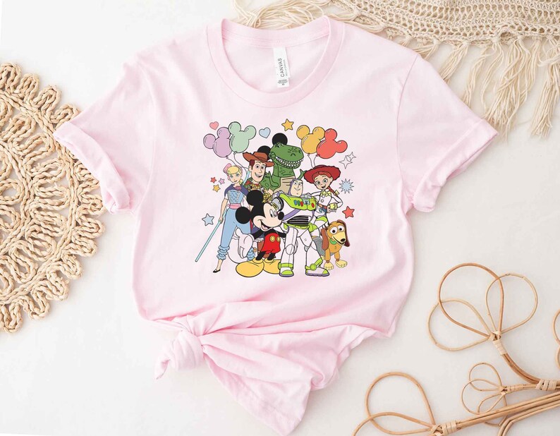 Puede incluir: Camiseta rosa con un estampado gr&aacute;fico de Mickey Mouse, Woody, Buzz Lightyear, Jessie, Rex y un extraterrestre verde de Toy Story. Los personajes est&aacute;n rodeados de estrellas y globos.