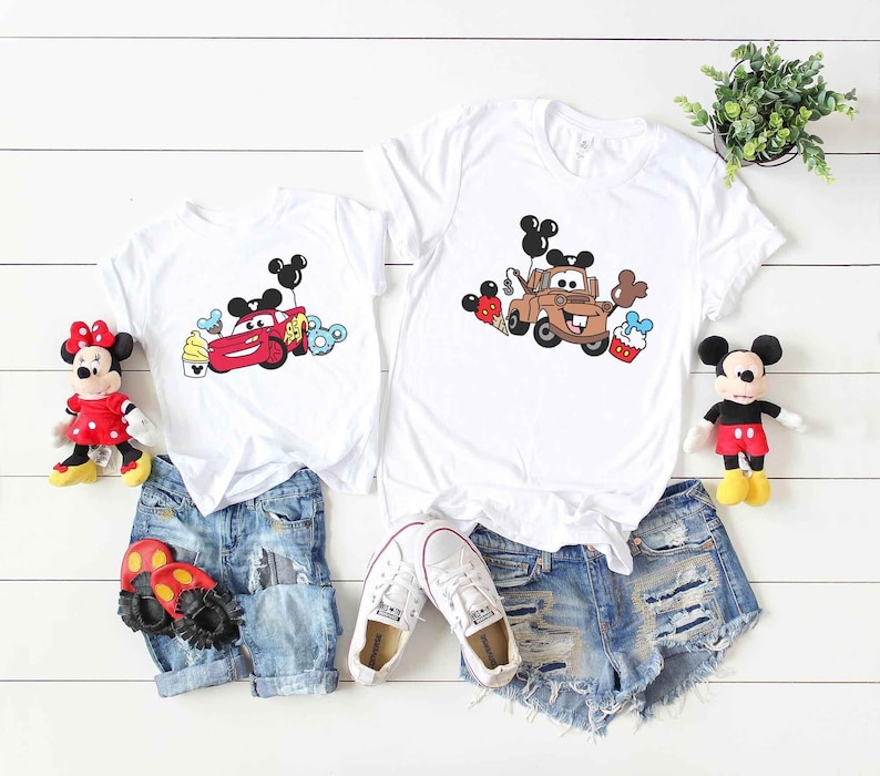 May include: Two white t-shirts with a cartoon image of a red car and a brown truck with Mickey Mouse ears and balloons. The shirts are paired with denim shorts and sneakers.