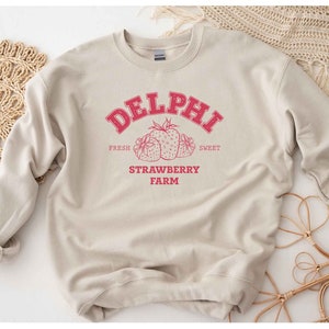 Op de afbeelding: Een beige sweatshirt met een rood grafisch ontwerp met de tekst "Delphi Strawberry Farm" met de woorden "Fresh" en "Sweet" aan weerszijden van het ontwerp. De grafiek bevat ook een cluster van rode aardbeien.