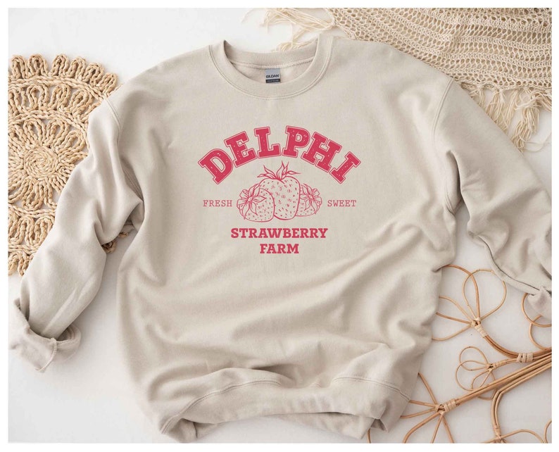 Op de afbeelding: Een beige sweatshirt met een rood grafisch ontwerp met de tekst "Delphi Strawberry Farm" met de woorden "Fresh" en "Sweet" aan weerszijden van het ontwerp. De grafiek bevat ook een cluster van rode aardbeien.