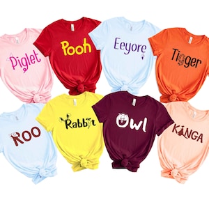 May include: Eight t-shirts featuring characters from Winnie the Pooh and The Jungle Book. The shirts are in various colors, including pink, red, blue, yellow, orange, maroon, and peach. Each shirt has a different character's name printed on it, including Piglet, Pooh, Eeyore, Tigger, Roo, Rabbit, Owl, and Kanga.