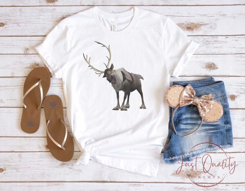 May include: White t-shirt with a cartoon image of a reindeer with antlers. The reindeer is brown and gray and has a surprised expression. The t-shirt is on a wooden surface with a pair of brown flip flops and a pair of denim shorts with a gold bow headband.
