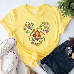 Puede incluir: Camiseta amarilla con una silueta de Mickey Mouse hecha de ilustraciones con tem&aacute;tica marina, incluyendo una sirena, conchas marinas y estrellas de mar.