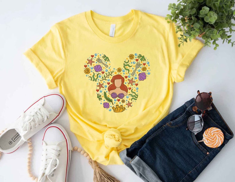 Puede incluir: Camiseta amarilla con una silueta de Mickey Mouse hecha de ilustraciones con tem&aacute;tica marina, incluyendo una sirena, conchas marinas y estrellas de mar.