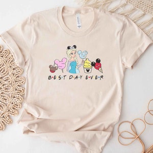 May include: A beige t-shirt with a graphic of a Disney princess character, a Mickey Mouse head, and various Disney treats. The text "BEST DAY EVER" is printed below the graphic.
