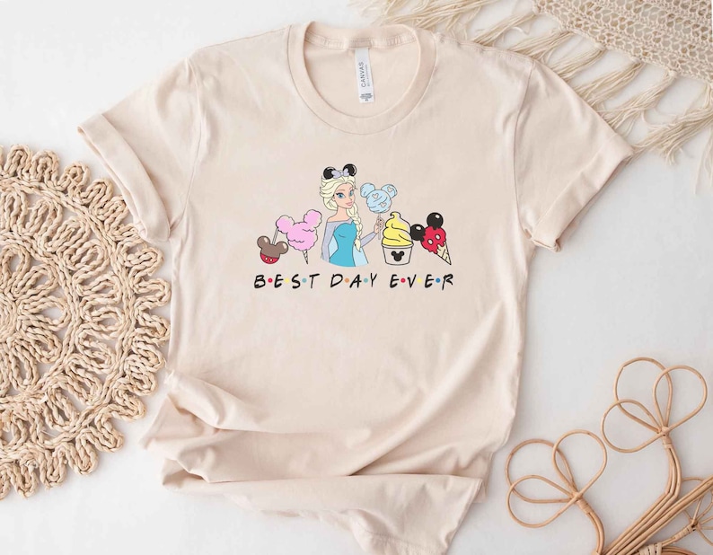 May include: A beige t-shirt with a graphic of a Disney princess character, a Mickey Mouse head, and various Disney treats. The text "BEST DAY EVER" is printed below the graphic.