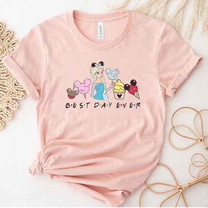 May include: A pink t-shirt with a graphic of a Disney princess holding a Mickey Mouse ice cream cone and other Disney treats. The text "BEST DAY EVER" is printed below the graphic.