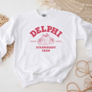 Op de afbeelding: Wit sweatshirt met een rood grafisch ontwerp met de tekst "Delphi Strawberry Farm" met een tekening van aardbeien. De tekst zegt "Fresh" en "Sweet" boven de aardbeien en "Strawberry Farm" eronder.