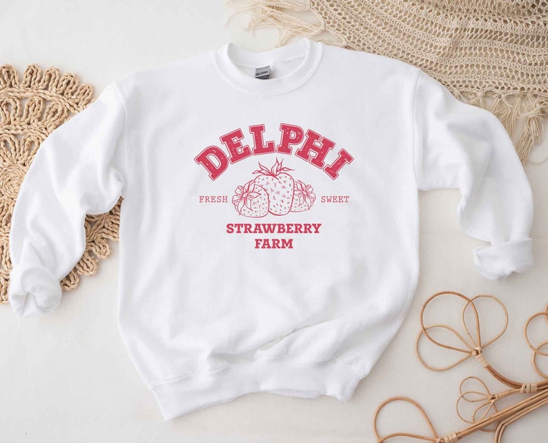 Op de afbeelding: Wit sweatshirt met een rood grafisch ontwerp met de tekst "Delphi Strawberry Farm" met een tekening van aardbeien. De tekst zegt "Fresh" en "Sweet" boven de aardbeien en "Strawberry Farm" eronder.
