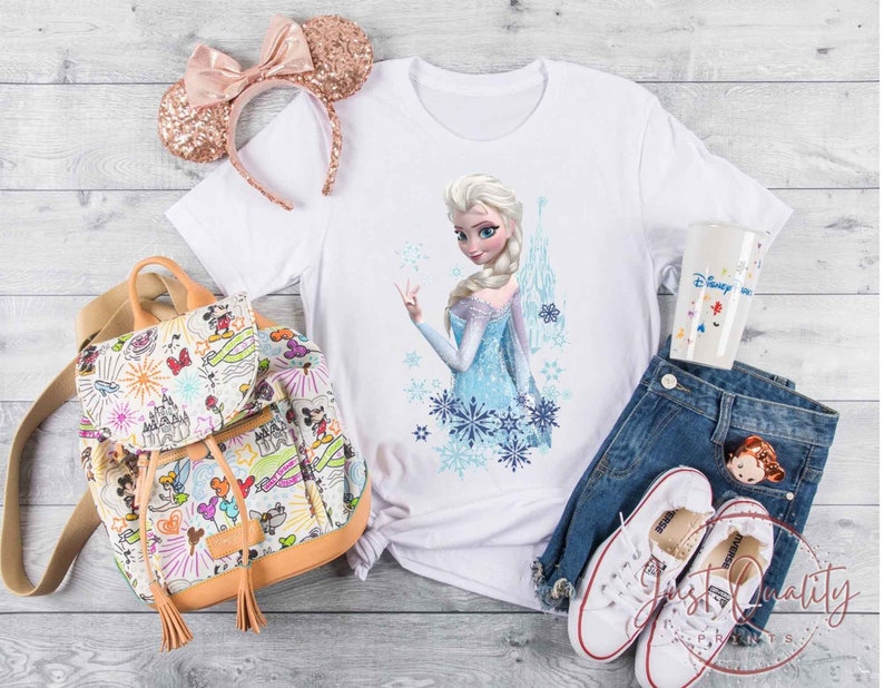 May include: A white t-shirt featuring a graphic of Elsa from Frozen, paired with blue jeans, white sneakers, and a Disney-themed backpack. A rose gold sequined Minnie Mouse ear headband and a Disney-themed tumbler are also present.