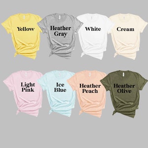 May include: A collection of unisex t-shirts in various colors, including yellow, heather gray, white, cream, light pink, ice blue, heather peach, and heather olive. The t-shirts are all folded and laid out on a gray background.