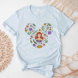 Puede incluir: Camiseta azul claro con un gr&aacute;fico de una cabeza de Mickey Mouse compuesta por varias im&aacute;genes de la pel&iacute;cula La Sirenita, incluyendo a Ariel, conchas marinas y algas.