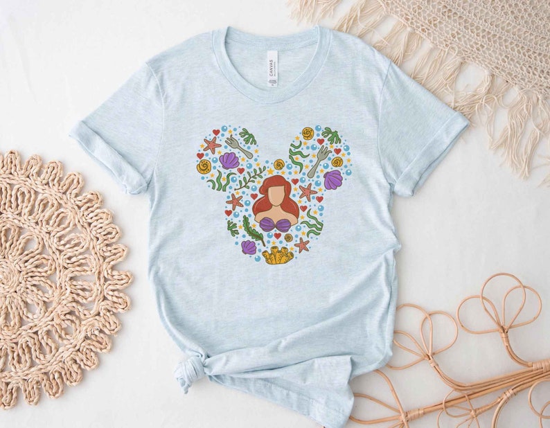 Puede incluir: Camiseta azul claro con un gr&aacute;fico de una cabeza de Mickey Mouse compuesta por varias im&aacute;genes de la pel&iacute;cula La Sirenita, incluyendo a Ariel, conchas marinas y algas.