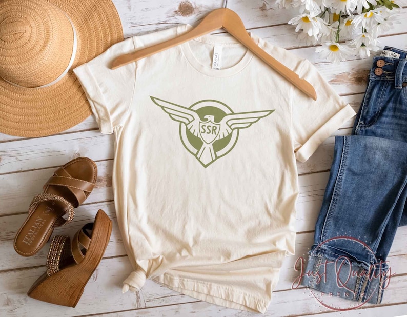 Op de afbeelding: Een beige T-shirt met een groene adelaar afbeelding en de tekst "SSR" in een cirkel. Het shirt ligt op een houten oppervlak met een strohoed, sandalen en een blauwe jeans.