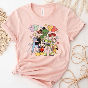 Puede incluir: Camiseta rosa con un estampado gr&aacute;fico de Mickey Mouse y personajes de Toy Story, incluyendo Woody, Buzz Lightyear, Jessie, Rex y Slinky Dog. Los personajes est&aacute;n rodeados de globos coloridos y estrellas.