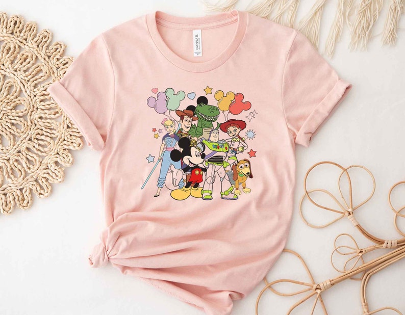 Puede incluir: Camiseta rosa con un estampado gr&aacute;fico de Mickey Mouse y personajes de Toy Story, incluyendo Woody, Buzz Lightyear, Jessie, Rex y Slinky Dog. Los personajes est&aacute;n rodeados de globos coloridos y estrellas.