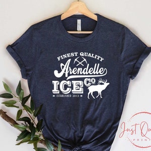 Puede incluir: Una camiseta azul marino con un dise&ntilde;o gr&aacute;fico blanco que dice "Finest Quality Arendelle Ice Co. Established 2013". El gr&aacute;fico tambi&eacute;n incluye un par de picos cruzados y una silueta de un alce.
