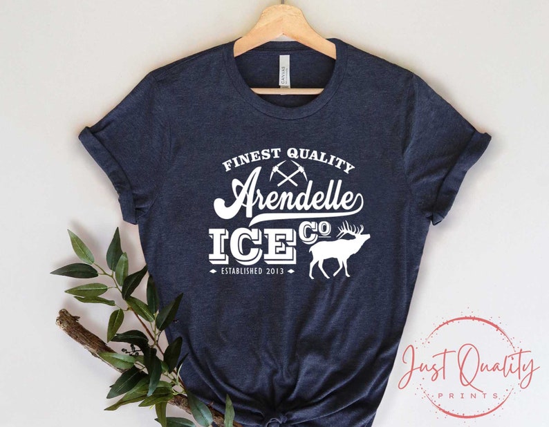 Puede incluir: Una camiseta azul marino con un dise&ntilde;o gr&aacute;fico blanco que dice "Finest Quality Arendelle Ice Co. Established 2013". El gr&aacute;fico tambi&eacute;n incluye un par de picos cruzados y una silueta de un alce.