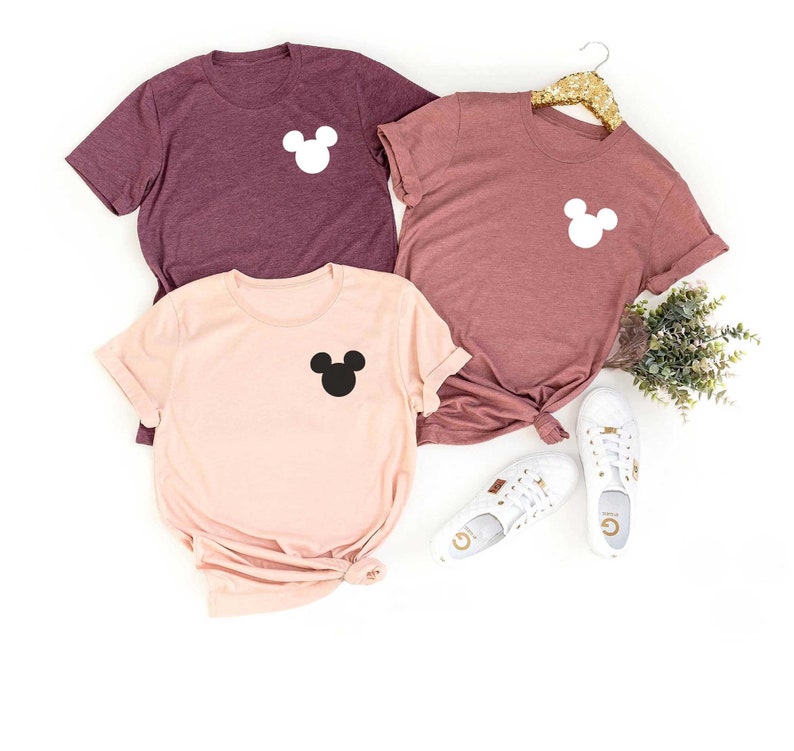 K&ouml;nnte beinhalten: Drei T-Shirts mit einer Mickey-Maus-Silhouette auf der Brusttasche. Die Shirts sind in den Farben Rosa und Rotbraun.