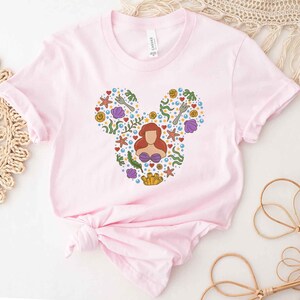 Puede incluir: Camiseta rosa con una silueta de Mickey Mouse compuesta por varias im&aacute;genes de La Sirenita, incluyendo a Ariel, conchas marinas y un tenedor.