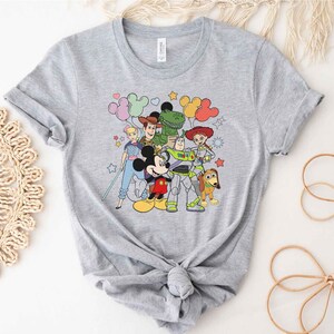 Puede incluir: Una camiseta gris con un estampado gr&aacute;fico colorido de Mickey Mouse, Woody, Buzz Lightyear, Jessie y Rex de Toy Story. Los personajes est&aacute;n rodeados de globos y estrellas.