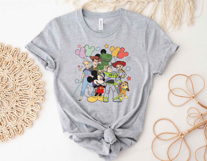 Puede incluir: Una camiseta gris con un estampado gr&aacute;fico colorido de Mickey Mouse, Woody, Buzz Lightyear, Jessie y Rex de Toy Story. Los personajes est&aacute;n rodeados de globos y estrellas.